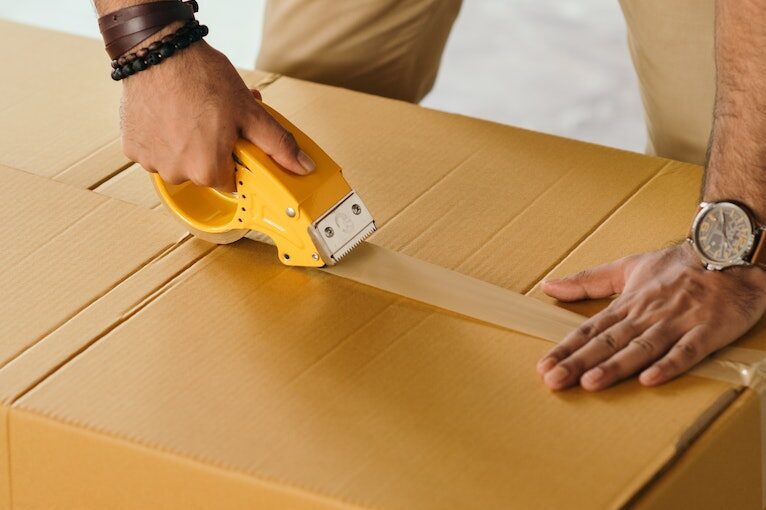 Unrecognizable man sealing box with duck tape while preparing for moving out